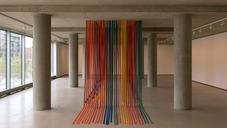 A colorful installation of hanging ribbons in red, orange, yellow, green, and blue cascades from the ceiling to the floor of a minimalist concrete gallery
