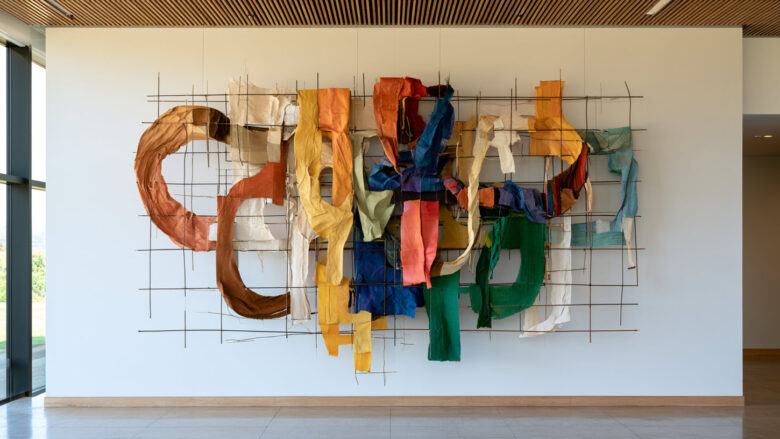 Abstract wall sculpture of torn colorful fabric draped across a black metal grid, evoking a commons of fragmented, interwoven contributions