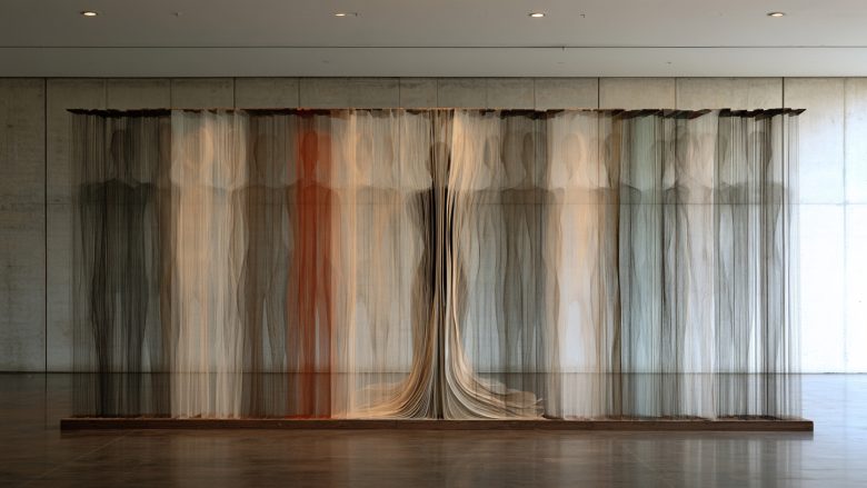 Suspended translucent human silhouettes in layered threads hanging in a concrete gallery, one in red